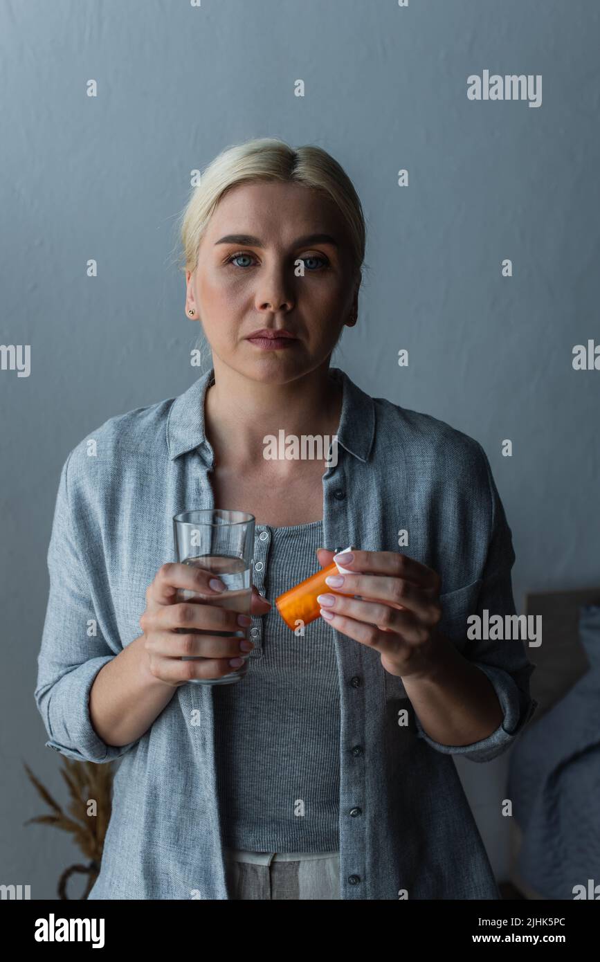blonde woman with menopause holding glass of water and bottle with medication Stock Photo