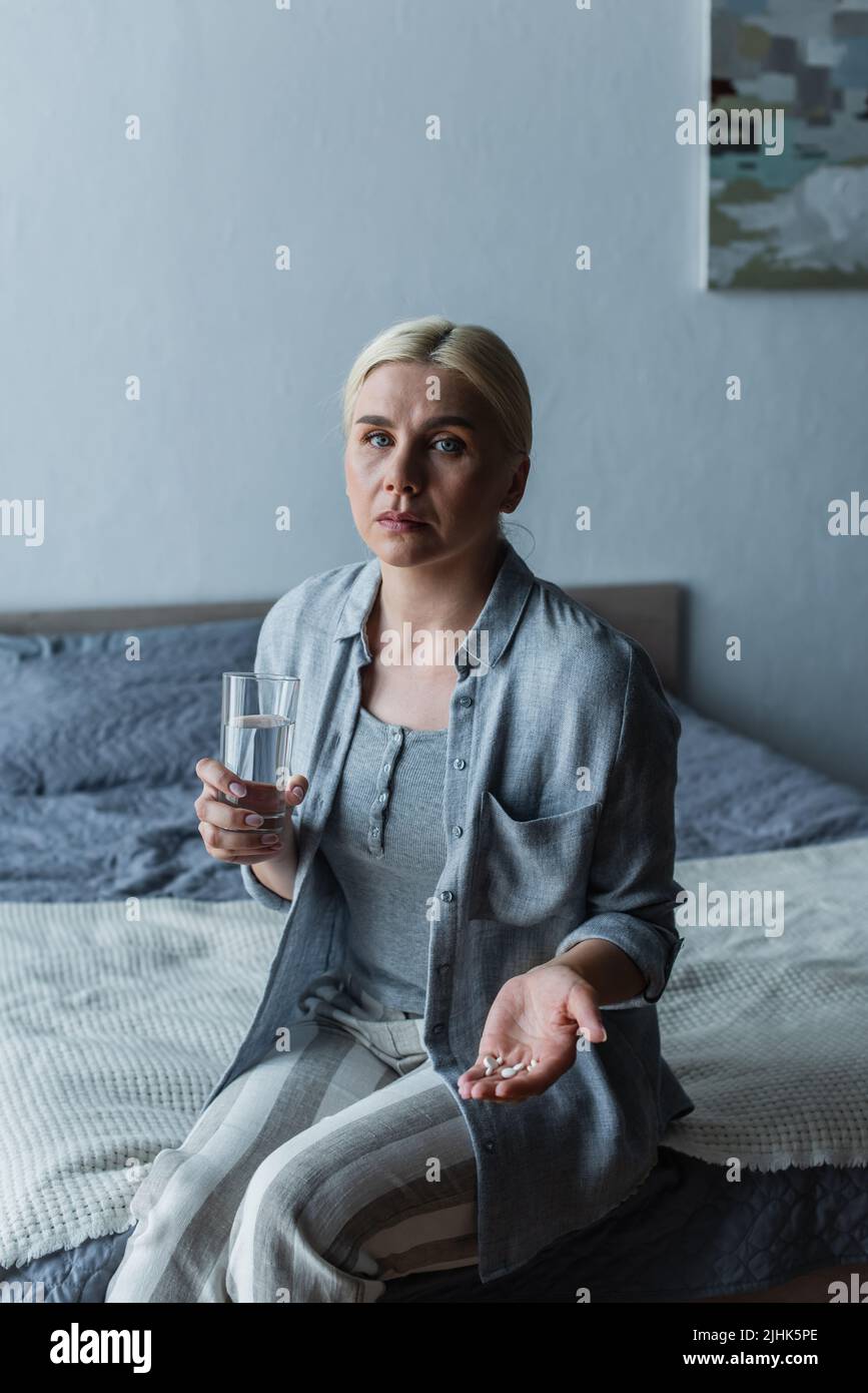 woman with menopause holding pills and glass of water while sitting on bed Stock Photo