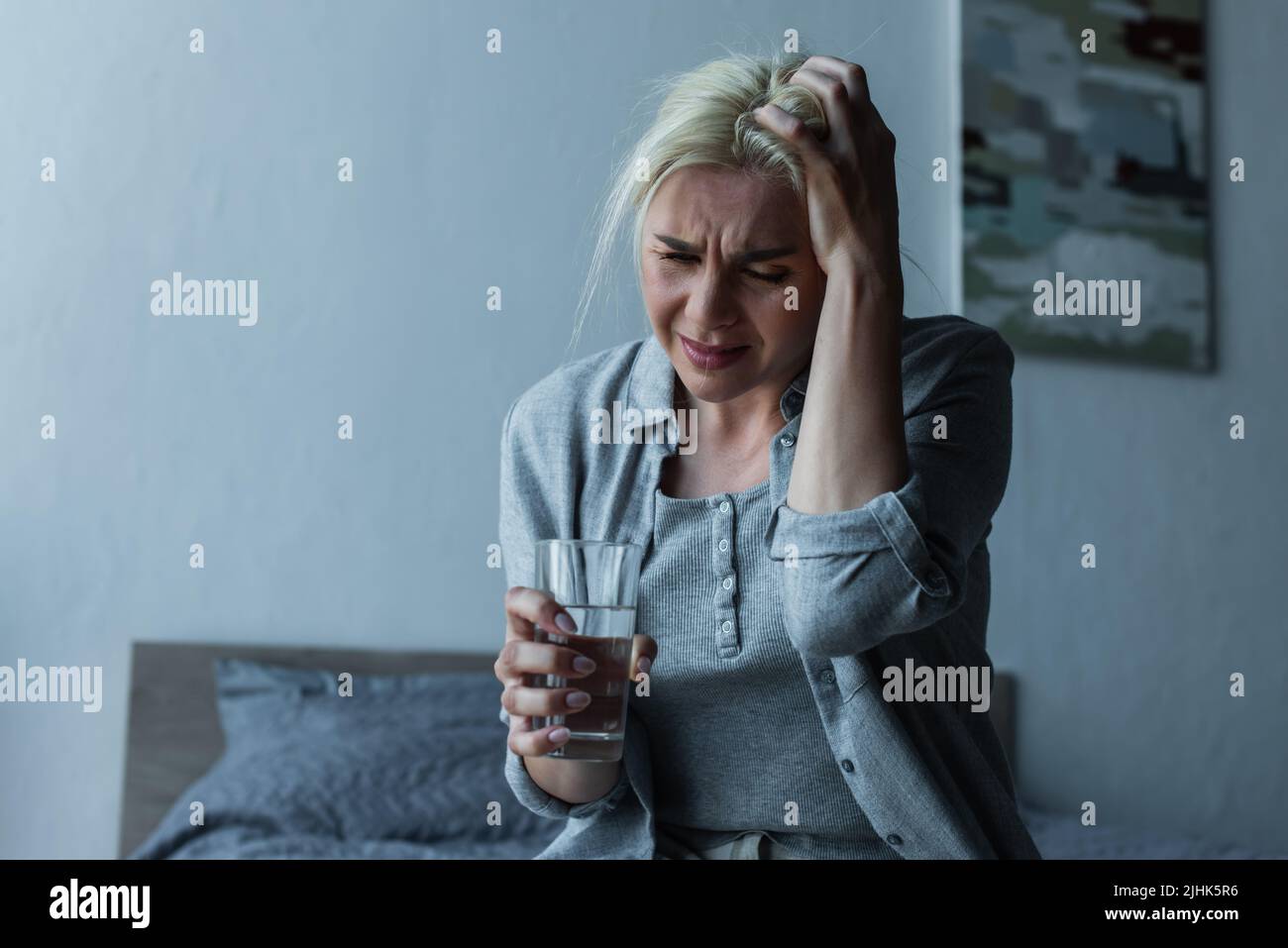 exhausted blonde woman holding glass of water while having migraine during menopause Stock Photo