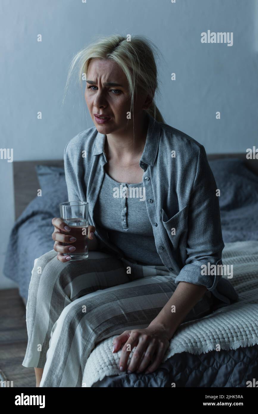 exhausted woman holding glass of water while sitting on bed during menopause Stock Photo