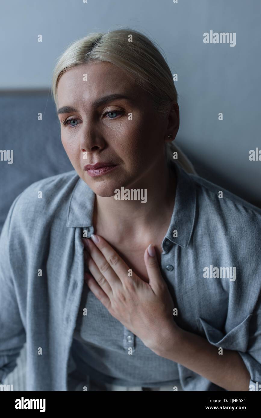 frustrated blonde woman with menopause touching chest in bedroom Stock