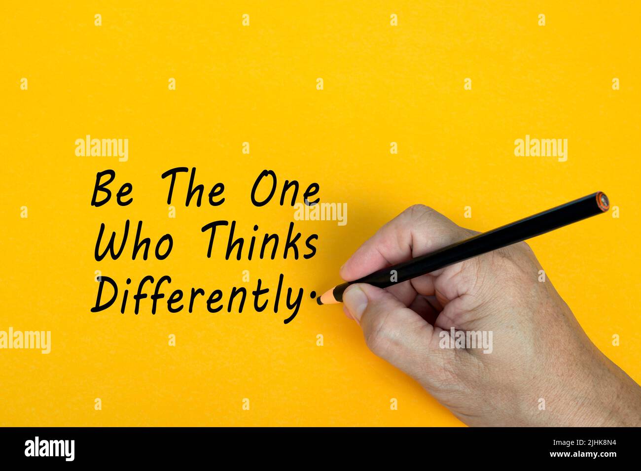 Male hand writes in black pencil the word BE THE ONE WHO THINKS DIFFERENTLY on a yellow background. Encouragement words. Stock Photo