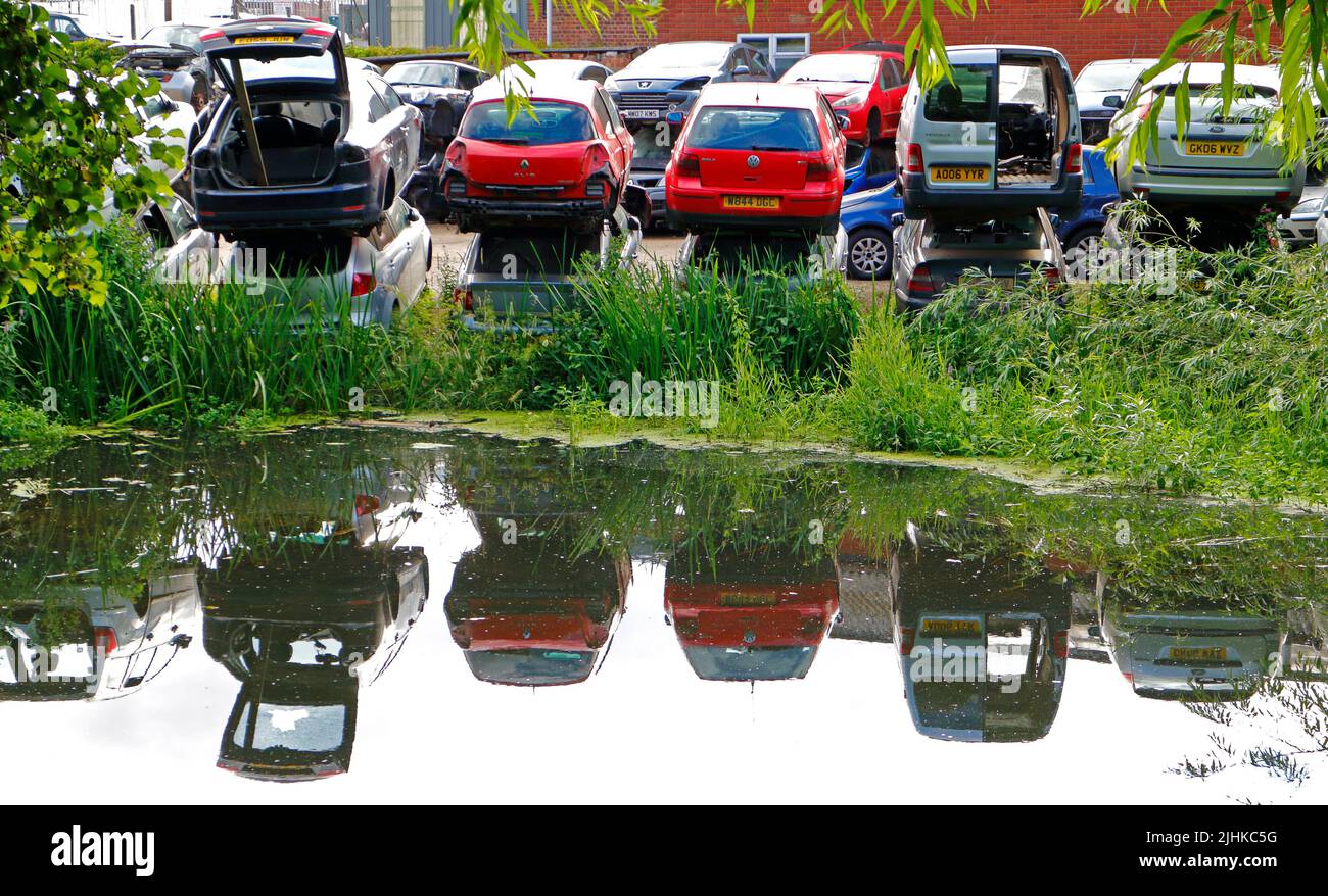 A view of stacked scrapped vehicles on the banks of the River Wensum with reflections in Norwich, Norfolk, England, United Kingdom. Stock Photo
