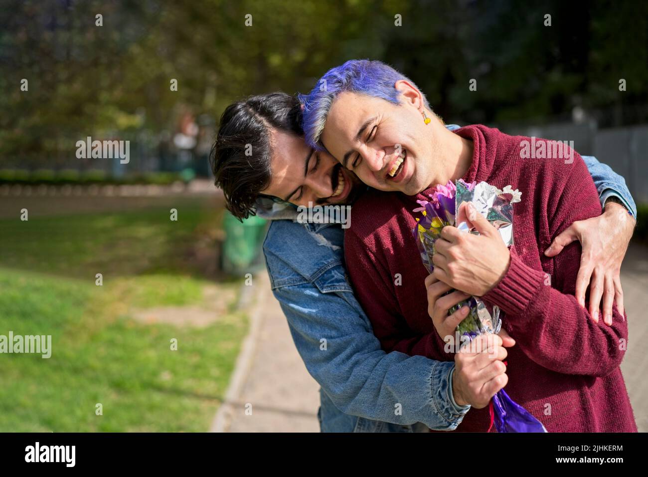 Couple of Latino gay men in a park embracing each other. One surprising the other with a bouquet of flowers as a gift Stock Photo