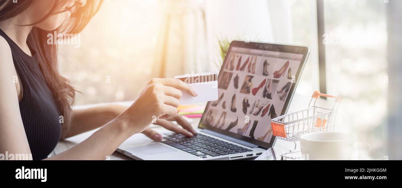 Horizontal online shopping banner, women looking for buying shoes in website on laptop and paying with credit cade on table at home. Stock Photo
