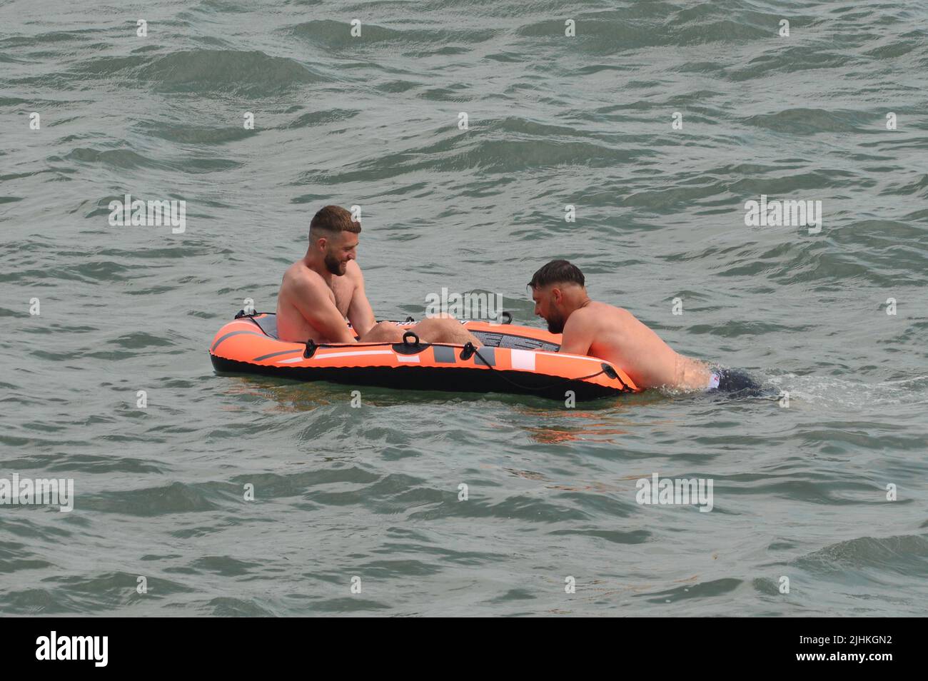 Boscombe, Bournemouth, Dorset, England, UK, 19th July 2022, Weather. The end is nigh for record breaking heatwave. Dark clouds and a rain shower over a busy beach in the afternoon. Two men in a dinghy stick to the plan. Credit: Paul Biggins/Alamy Live News Stock Photo