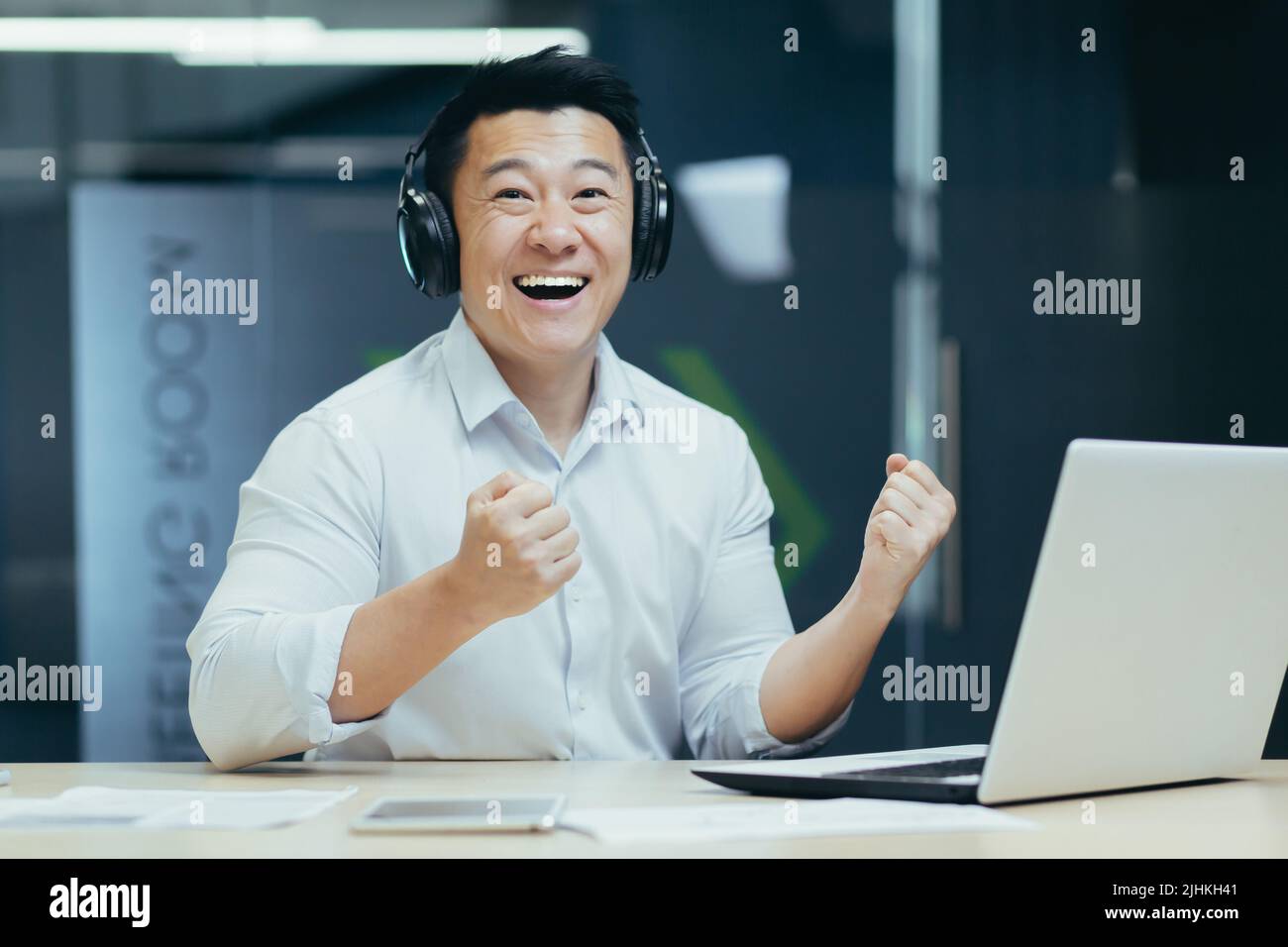 portrait of businessman with headphones, asian man smiling and looking at camera, boss watching sports match at workplace Stock Photo