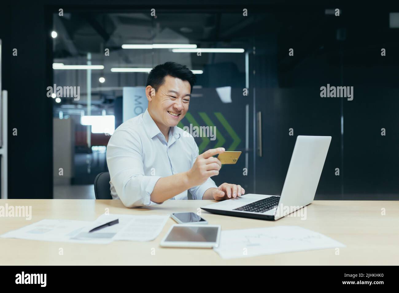 Happy Asian businessman working in modern office, man making online money transfer, using laptop and holding bank credit card Stock Photo