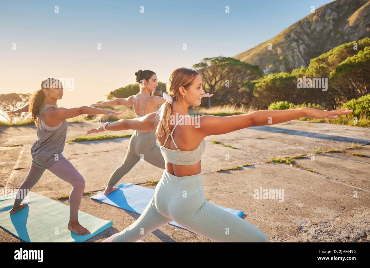 Diverse smiling yoga women in warrior pose during outdoor practice in remote nature. Group of happy active friends using mat and balancing while Stock Photo