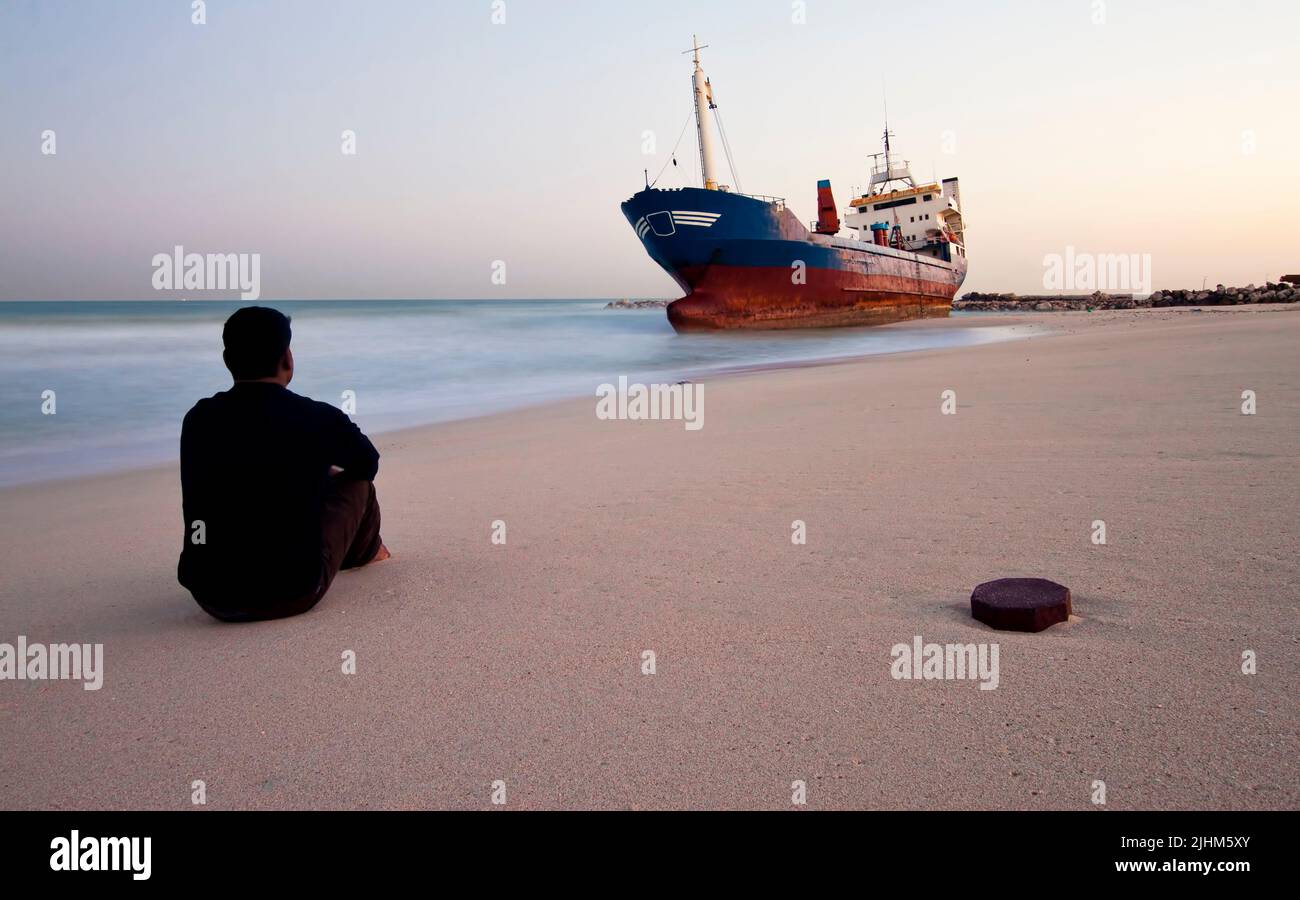 Middle aged man sitting on the beach and watching the shored ship in Sharjah, United Arab Emirates Stock Photo