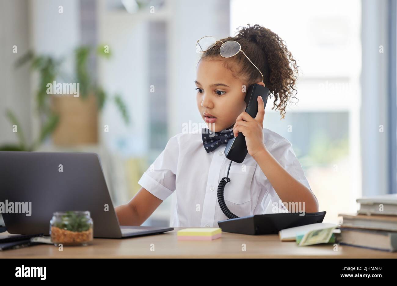 Let me find that for you. an adorable little girl dressed as a businessperson sitting in an office and using a laptop and telephone. Stock Photo