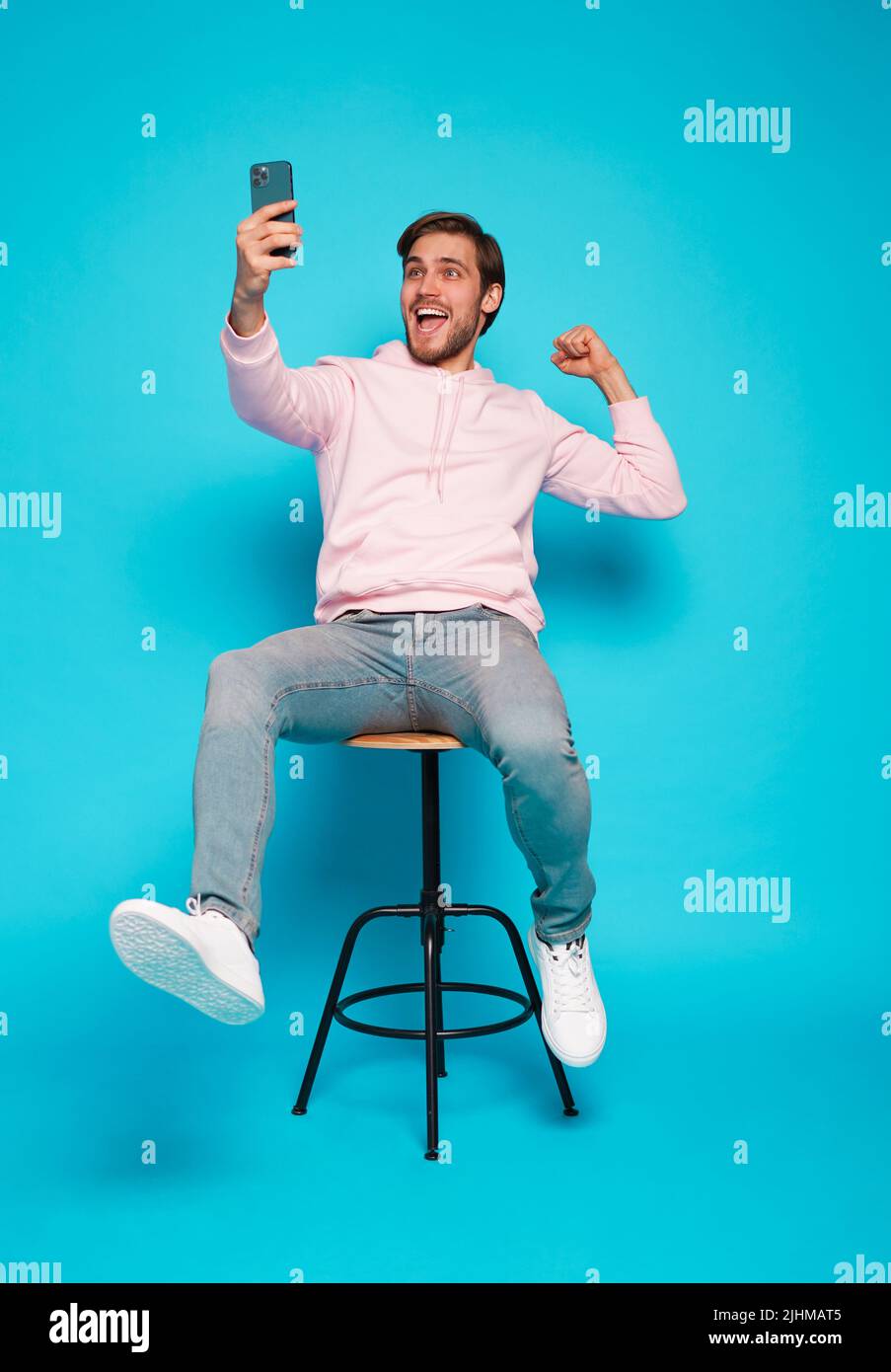 Online Profit. Portrait of a joyful young man sitting on chair and holding mobile phone isolated over light blue background, celebrating financial suc Stock Photo