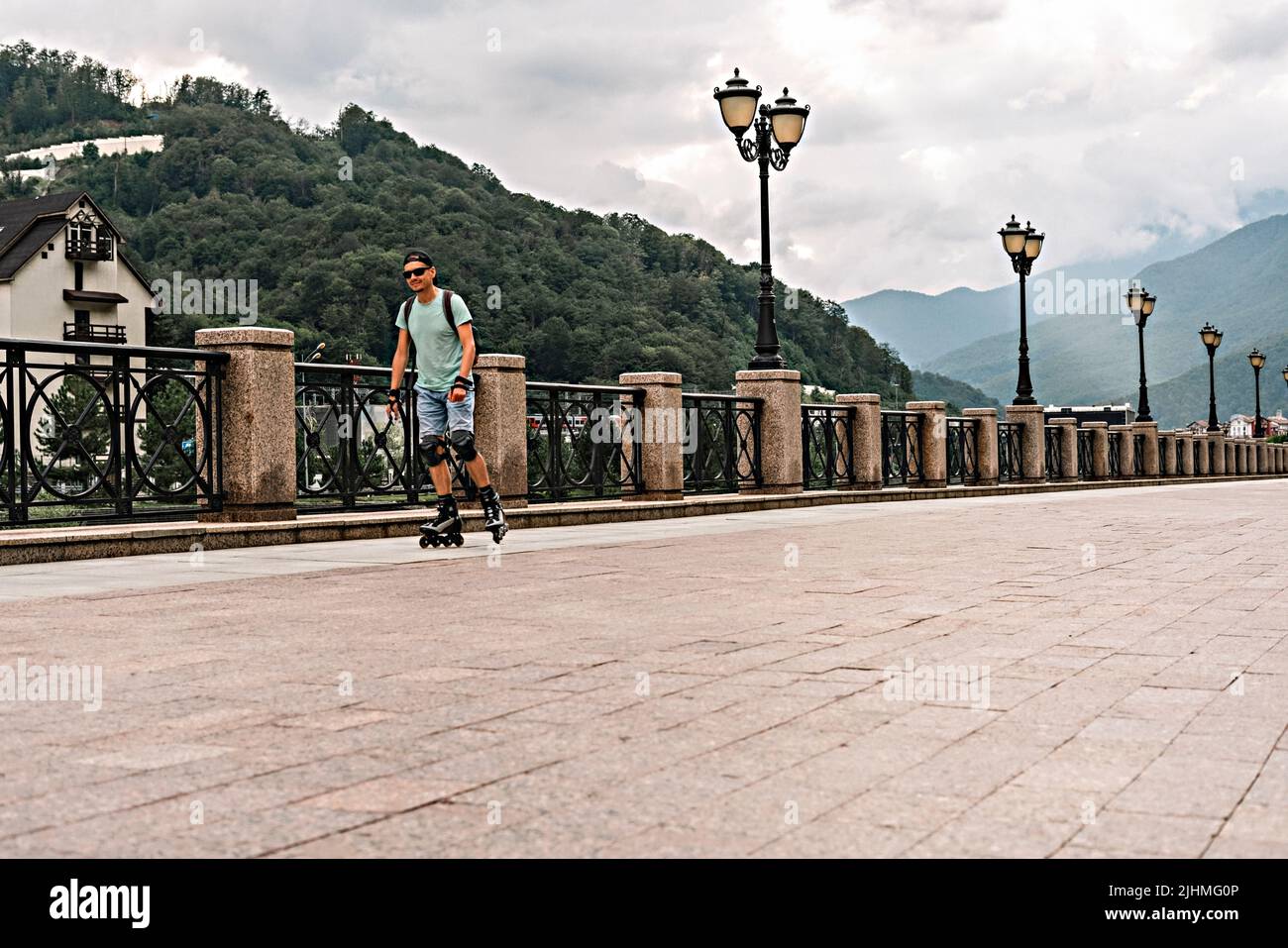 Young man in blue clothes in protective equipment riding on roller skates along the embankment against the background of forest and mountains in summe Stock Photo