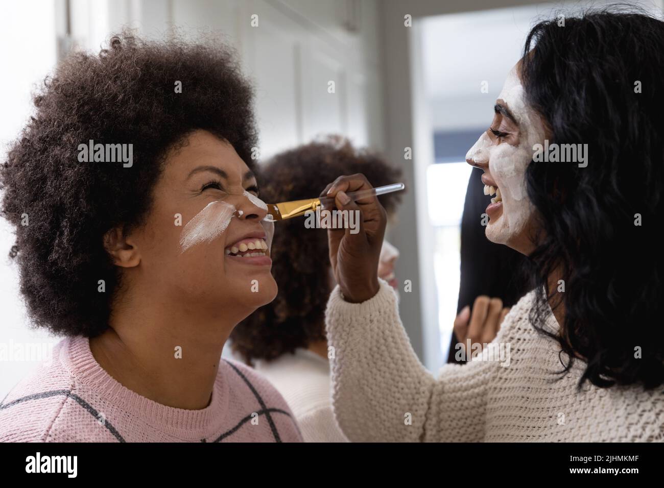 Happy biracial young woman with beauty cream on face applying face pack to female friend at home Stock Photo