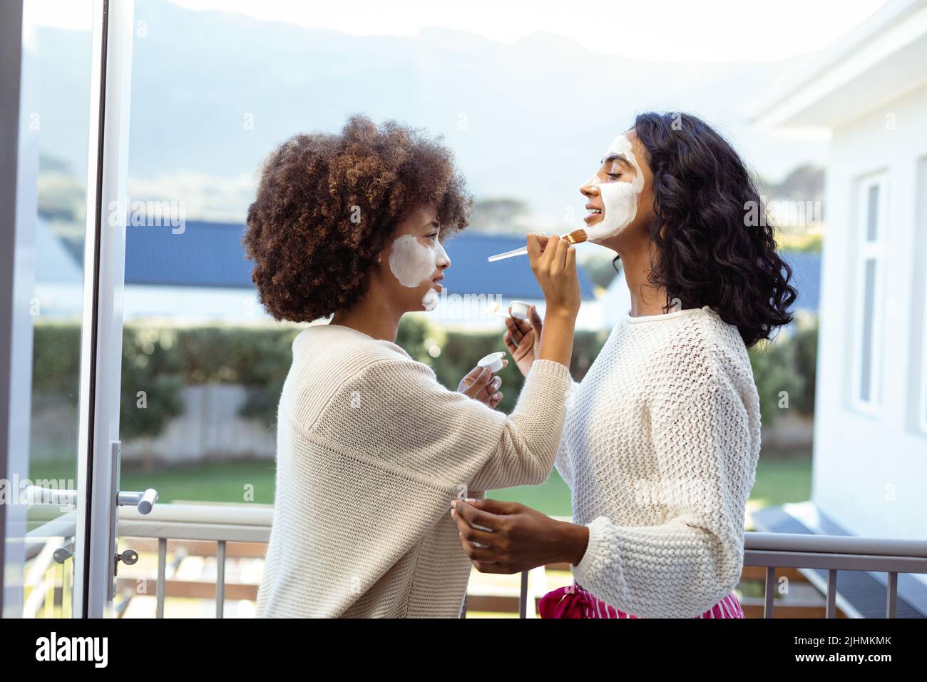 Side view of biracial female friends applying beauty cream on each others faces in balcony Stock Photo