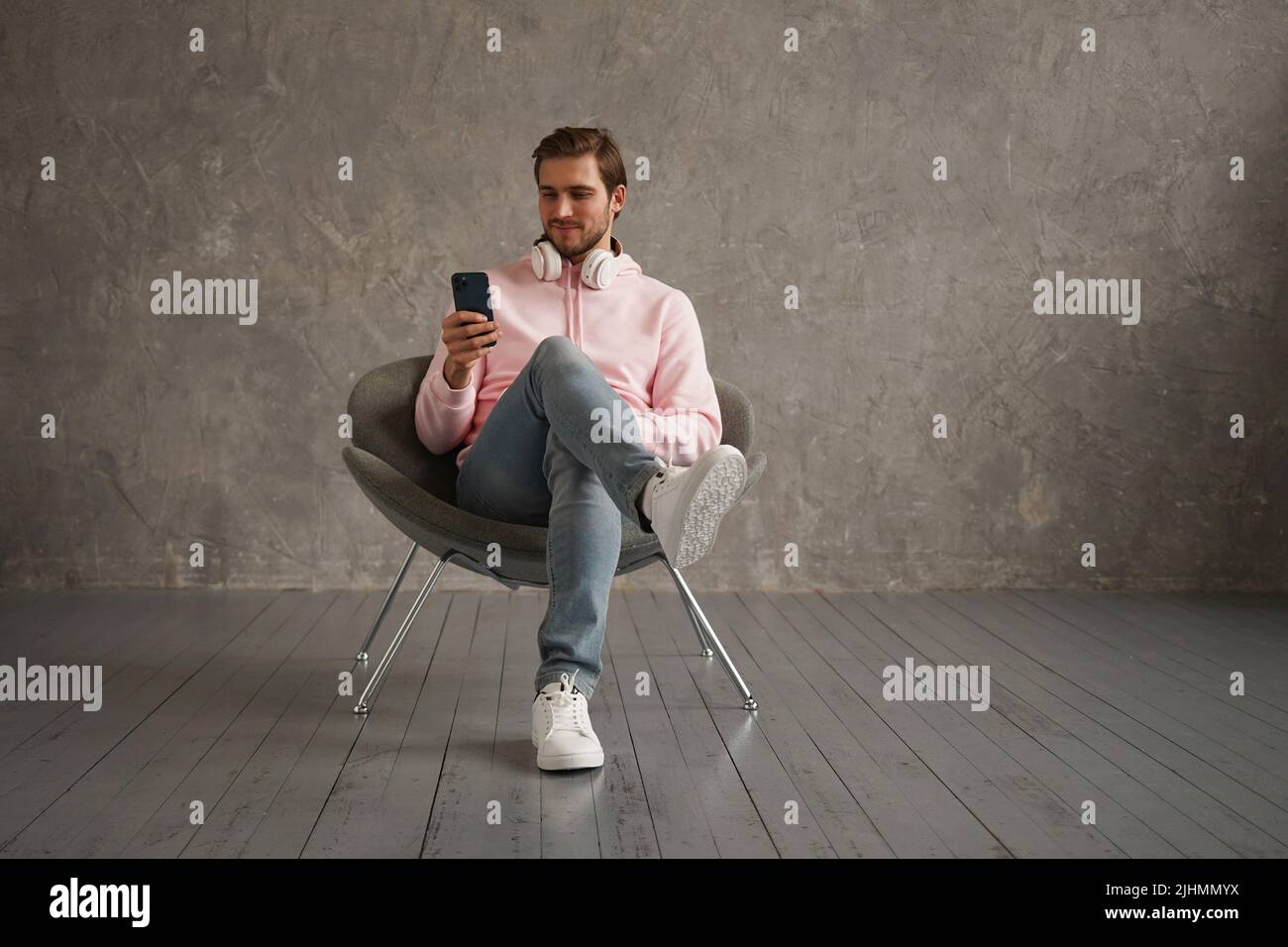 Cheerful Guy Using Mobile Phone With New Application Sitting In Armchair Over Gray Wall Background Stock Photo