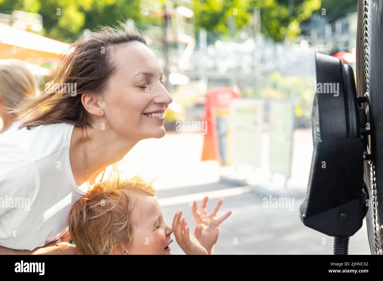 Profile side close-up view young adult woman with son enjoy fresh moisturized air blowing from big cooling fan machine hot sunny summer weather Stock Photo