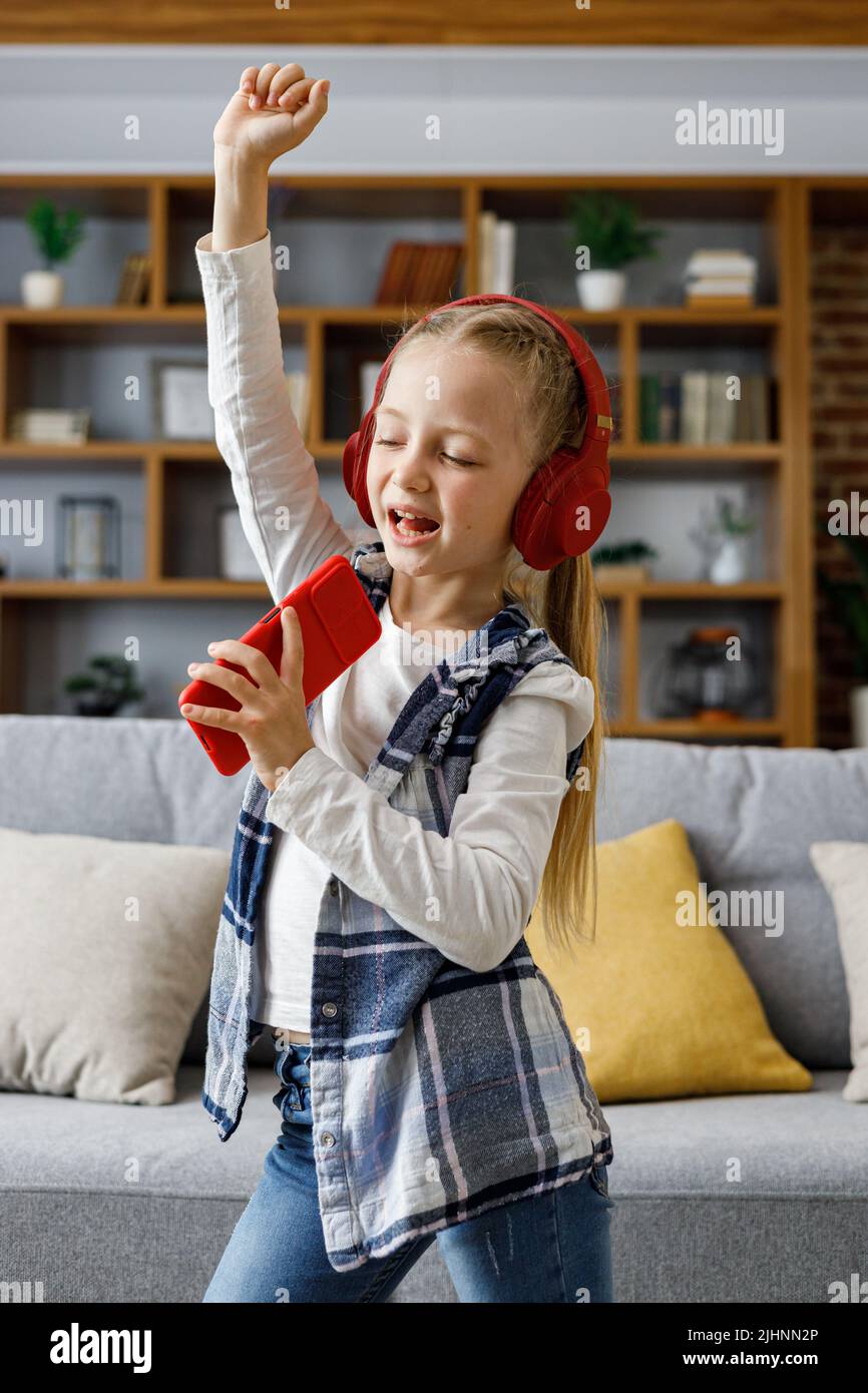 Happy little girl wearing red headphones dancing and singing using smartphone like microphone. Cute child enjoy listening favorite music tracks in Stock Photo