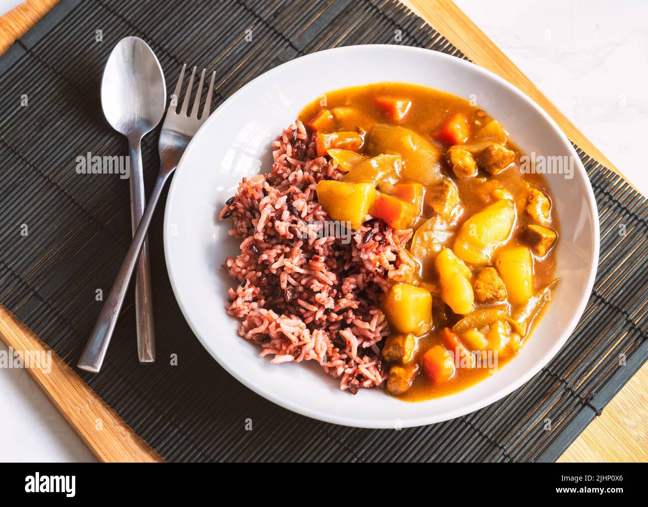 Japanese curry with rice berry in round white plate, healthy food with natural light, Japanese curry and rice with spoon and fork black wooden placema Stock Photo