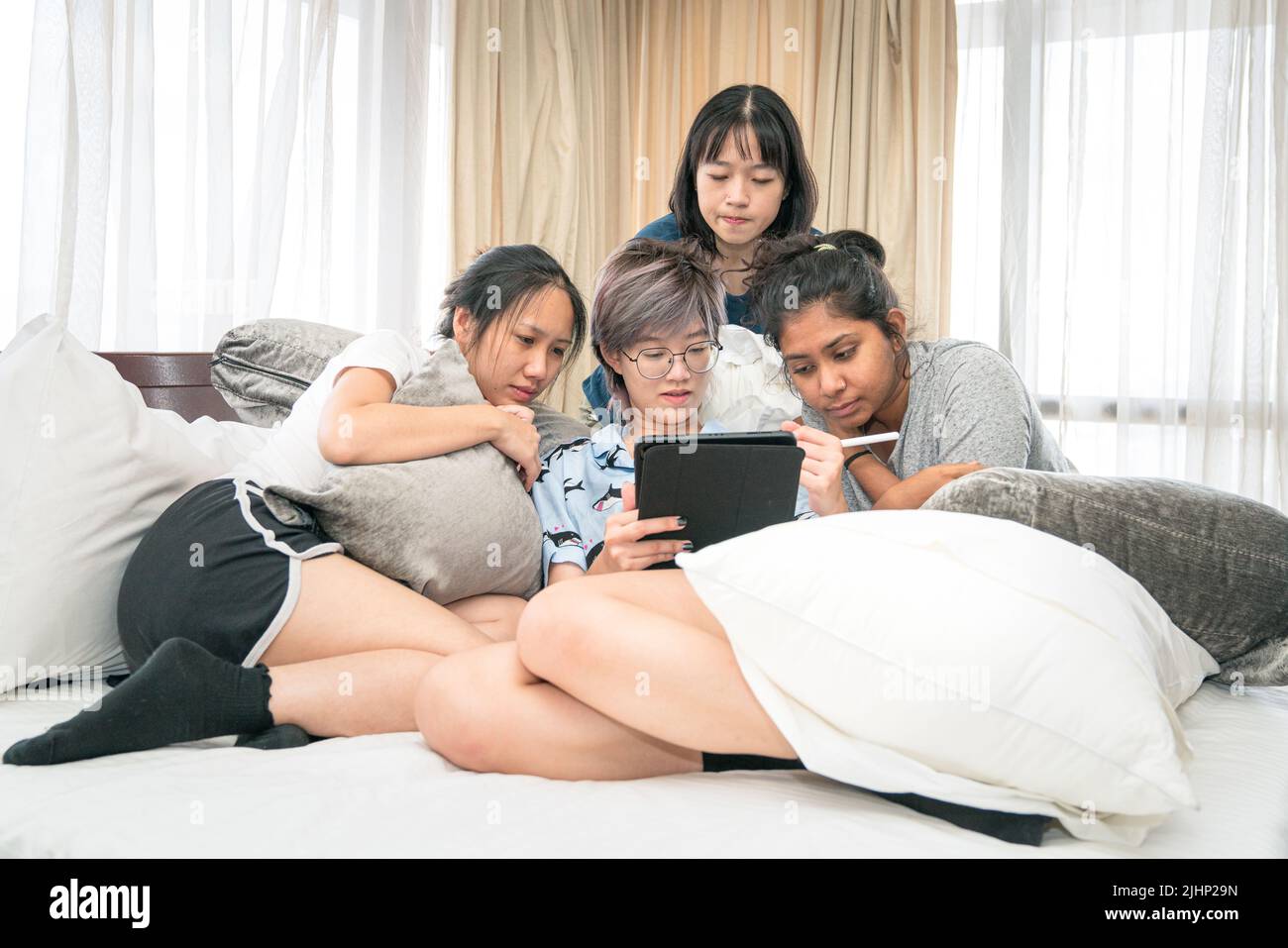 Young multi-ethnic women lying on bed browsing digtial tablet together. Digital lifestyle or sleepover concept. Stock Photo