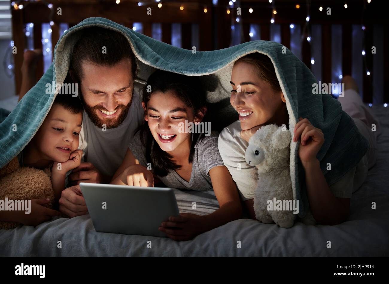 Happy caucasian family with two children using digital tablet lying under blanket in the dark at night with their faces illuminated by device screen Stock Photo