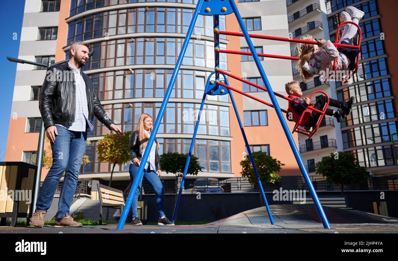 Happy family - father, mother and children having fun together on playground in new city district. Parents swinging daughter and son. Modern residential buildings on background. Stock Photo