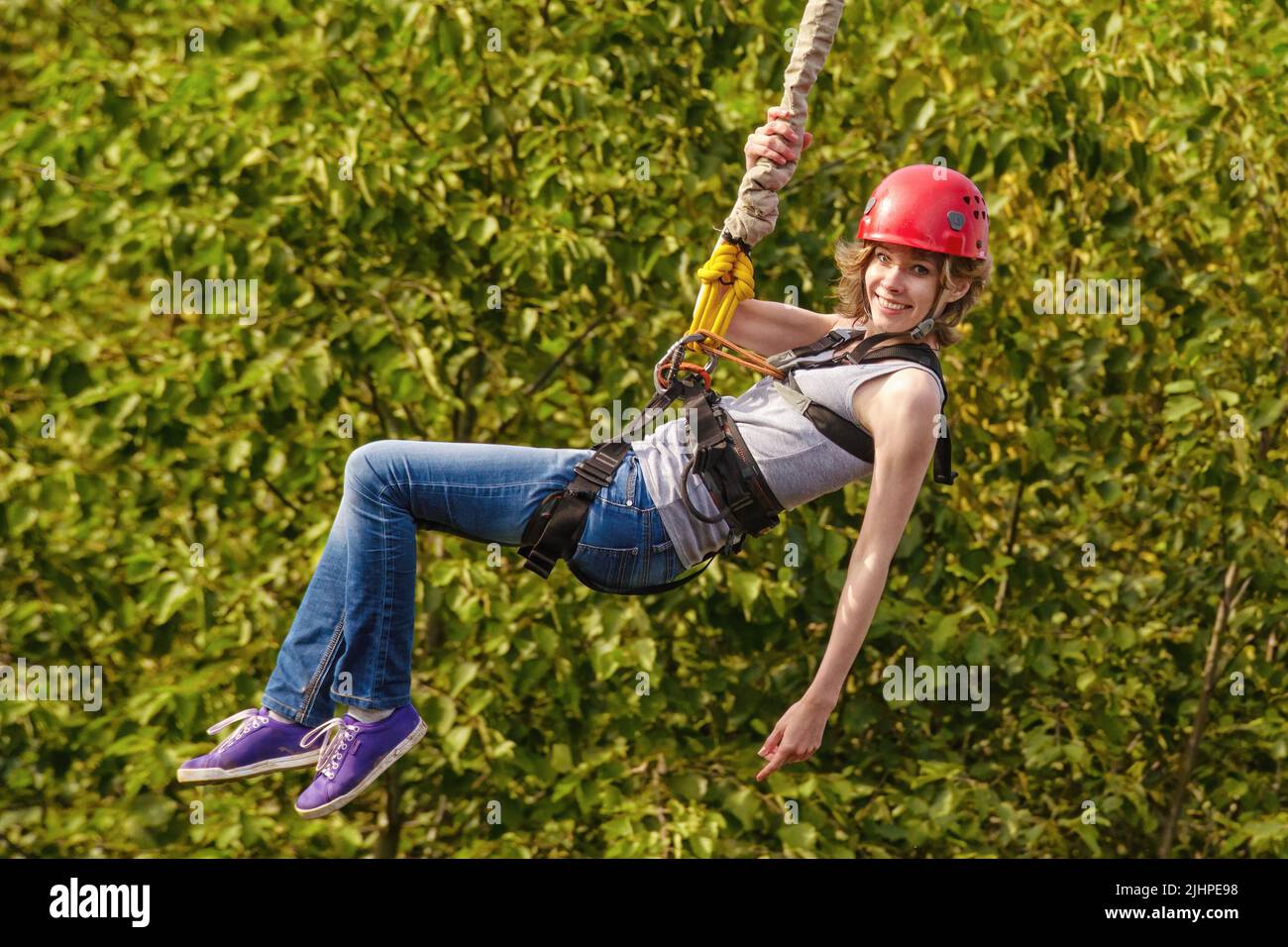 Beautiful young woman in a helmet hanging on a rope after the bungee jump against green foliage Stock Photo