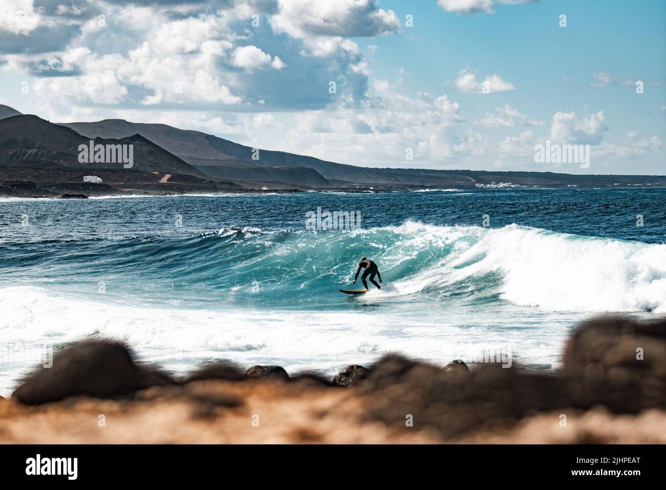 Wild rocky coastline of surf spot La Santa Lanzarote, Canary Islands, Spain. Surfer riding a big wave in rocky bay, volcano mountain in background. Stock Photo