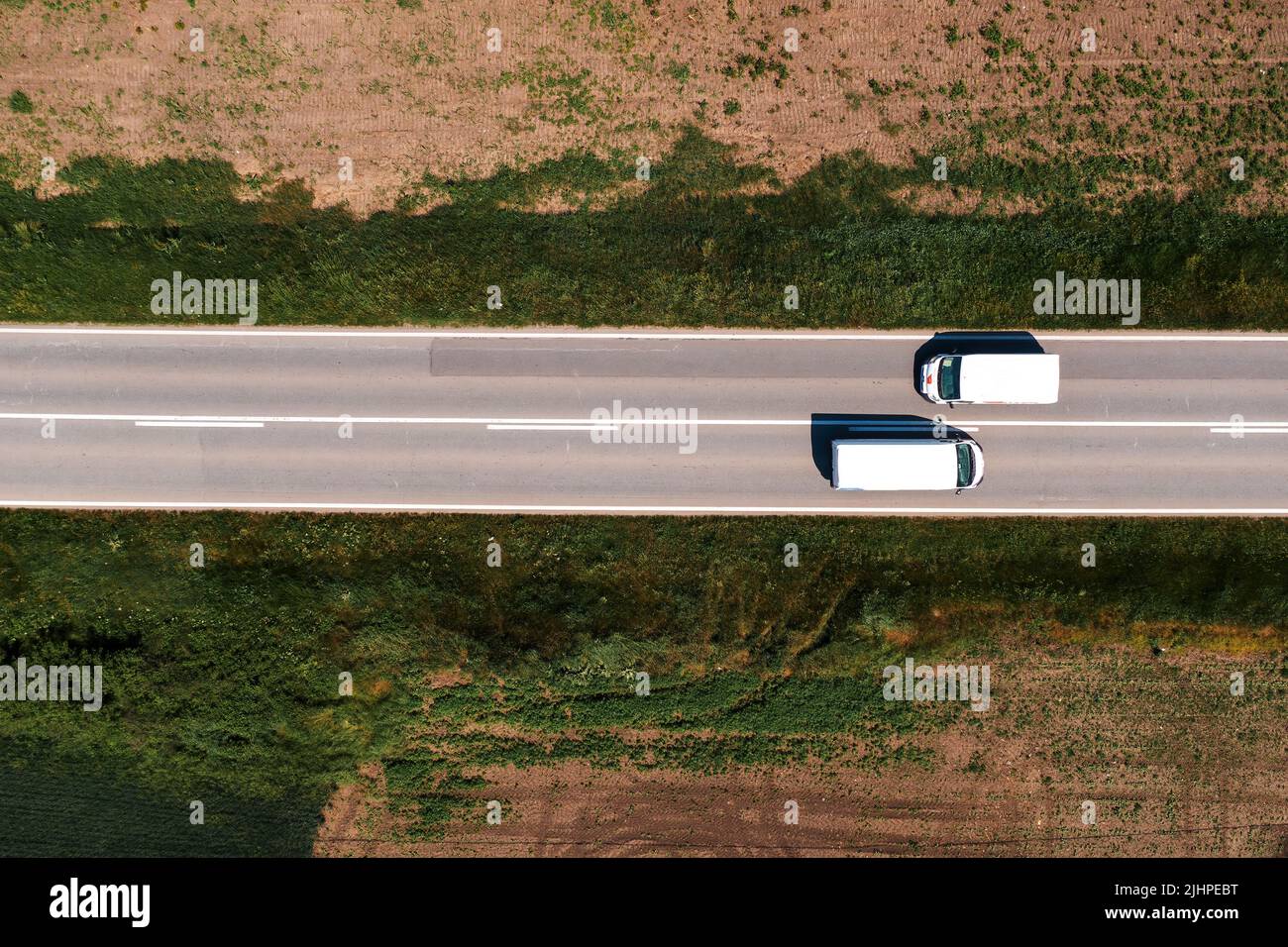 Aerial shot of two white van vehicles passing by each other on highway through countryside landscape on sunny summer day, directly above Stock Photo