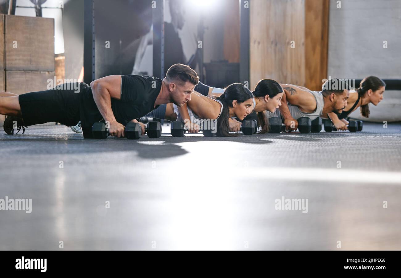 Diverse group of active young people doing push up exercises with dumbbells while training together in a gym. Focused athletes doing press ups with Stock Photo