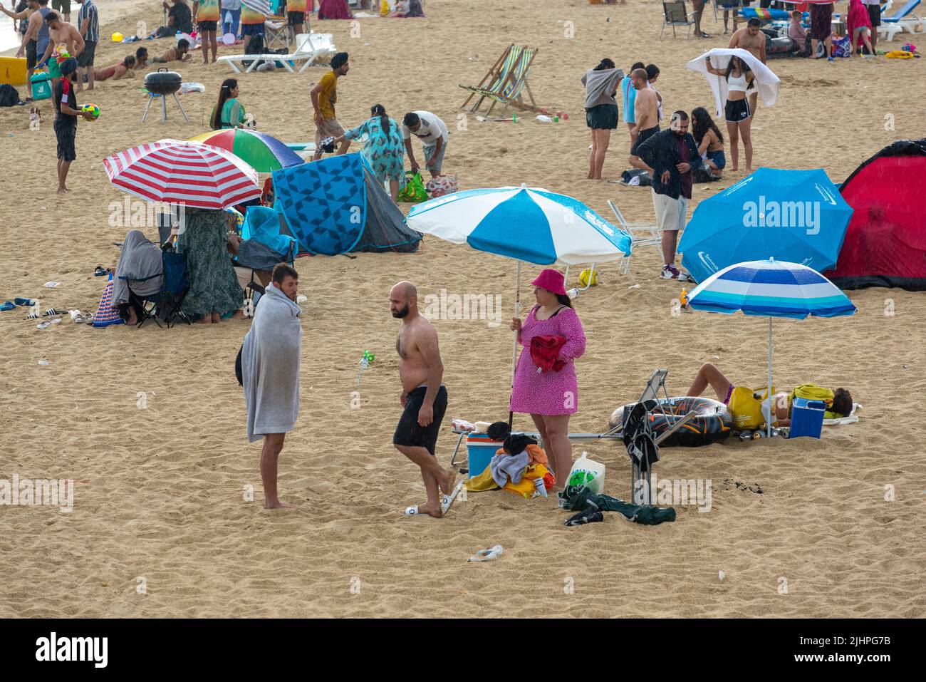 Boscombe, Bournemouth, Dorset, England, UK, 19th July 2022, Weather. People sitting on beach using sun umbrellas as rain umbrellas in heatwave shower. Stock Photo