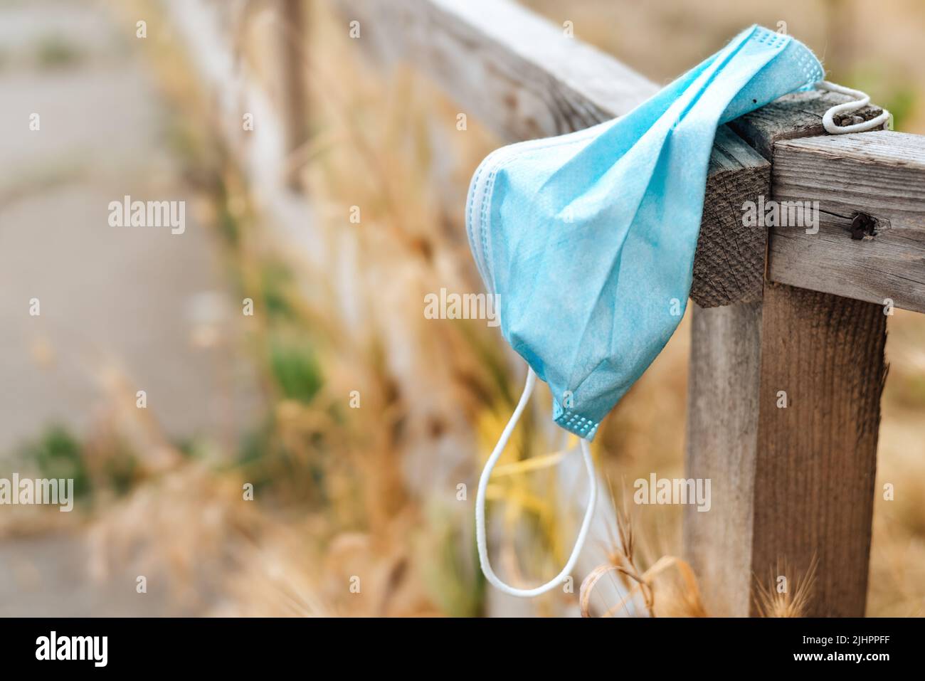 Waste during COVID-19. A blue medical face mask hangs on a wooden fence and is thrown into the nature among the yellow grass. End of quarantine. Environmental pollution with plastic. Stock Photo