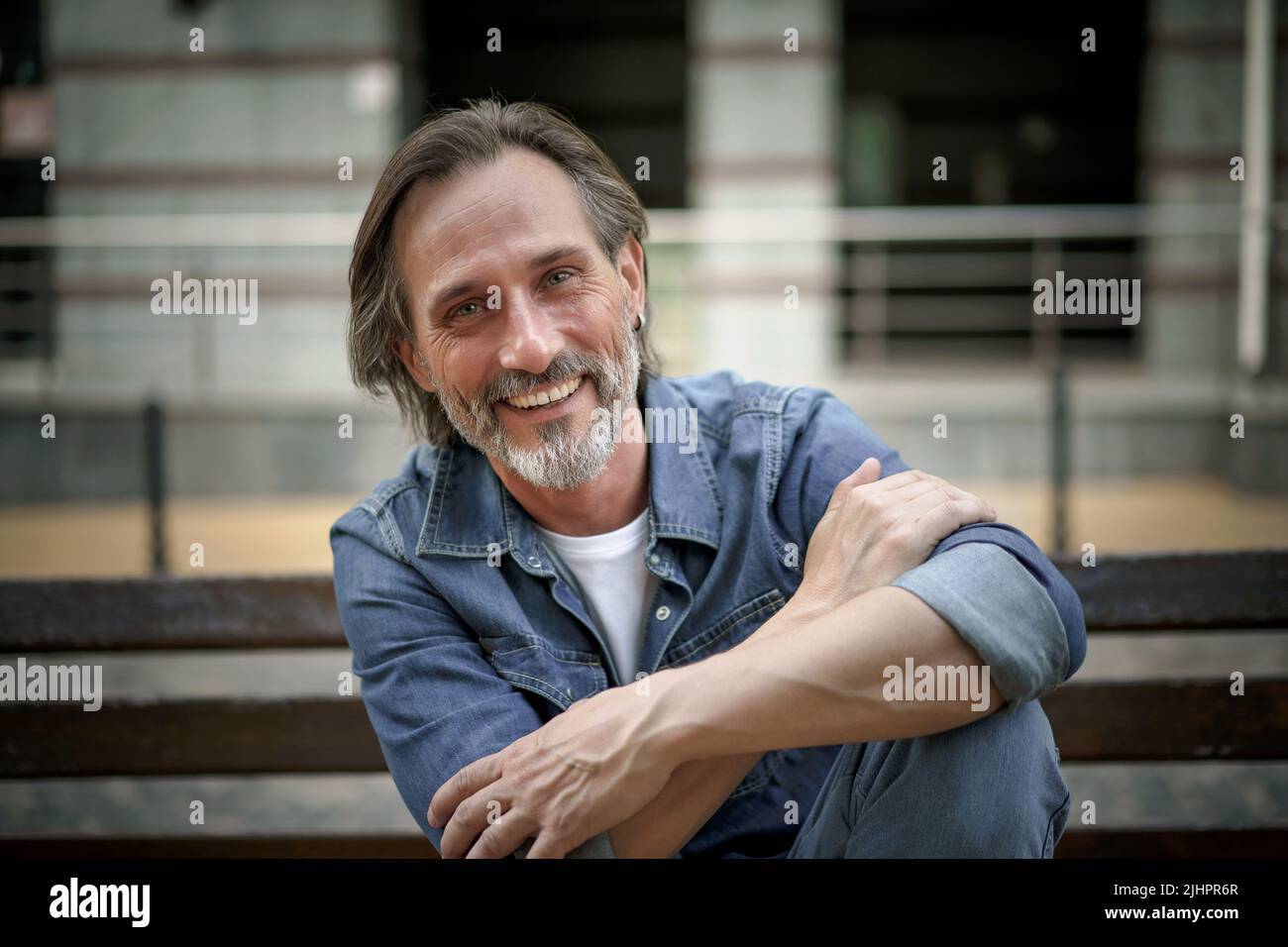 Smiling handsome middle aged man with grey beard sits on the bench with hands folded leaned on one lifted foot outdoor at urban city background enjoying free time. Travel and people concept.  Stock Photo