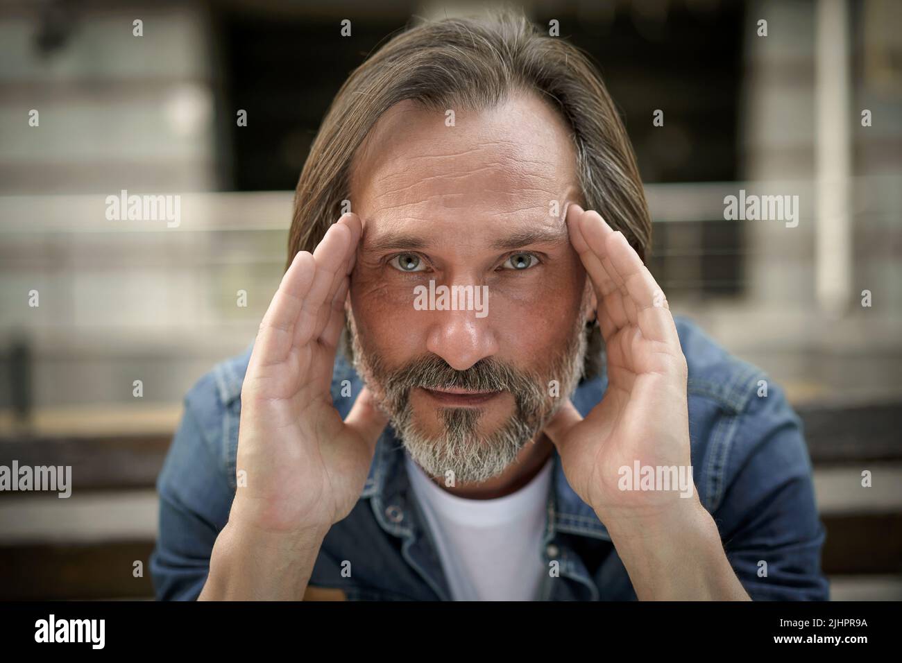 Playful middle age man with grey beard deep in his thoughts with positive emotions on his face. Happy thinking of life middle aged man looking at camera sitting on the bench in old town. Stock Photo