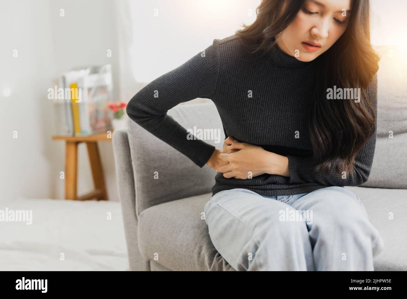 Young woman suffering from strong abdominal pain while sitting on sofa at home Stock Photo