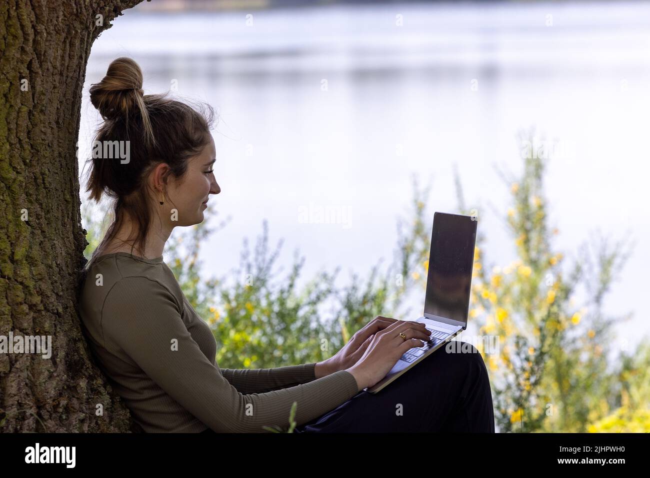 Work from anywhere. Remote freelancer work in nature using renewable energy via a foldable solar panel. Young woman, female freelancer working with laptop with Beautiful view of forest and lake. freelancer paradise, freedom of teleworking. distance. High quality photo Stock Photo