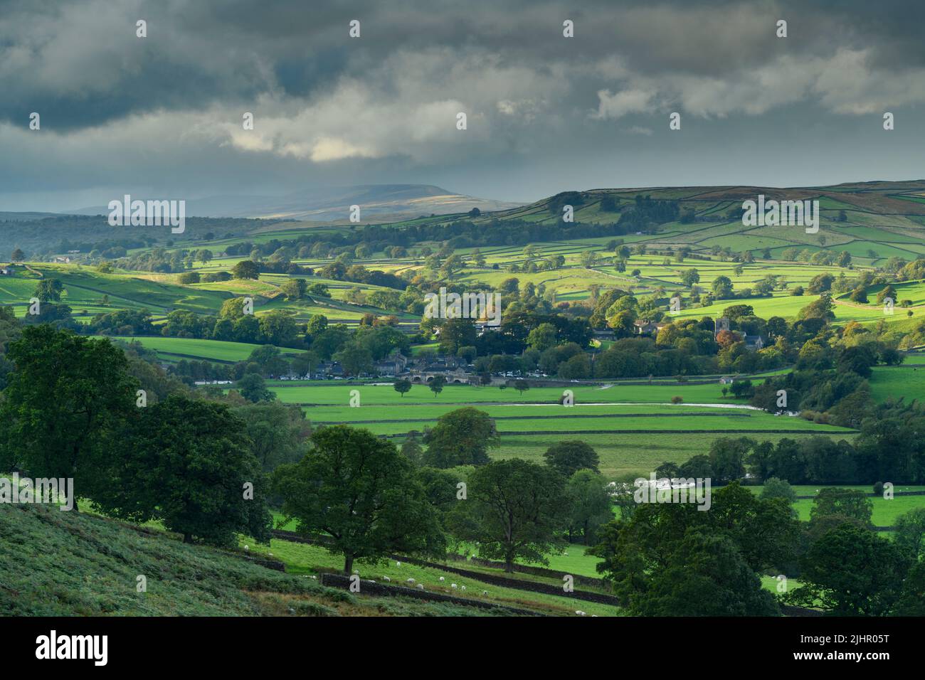 Scenic Burnsall in valley (hillside slopes, high uplands, green fields & pastures, dry-stone walls, cloudy sky & sun) - Upper Wharfedale, England, UK. Stock Photo