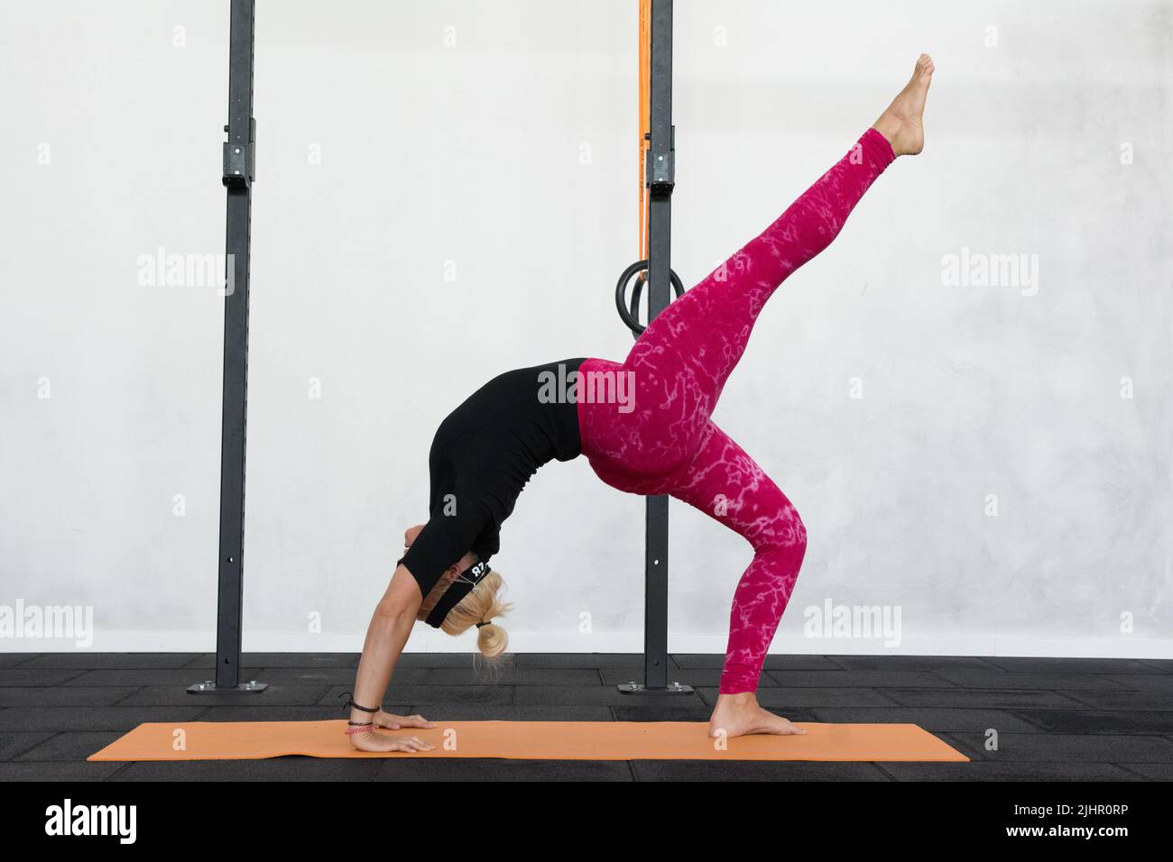 Youth blonde woman performing ekapada chakrasana, advanced back bending with right leg upwards yoga pose wearing colorful pink yoga leggings. Stock Photo