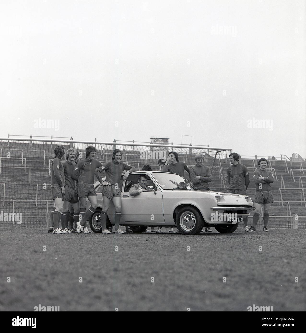 1974, historical, football manager, Malcolm Allison sitting in 2-door Vauxhall Chevette hatchback car on the pitch at Selhurst Park, with team players standing around. Selhurst Park is the home of Crystal Palace Football Club. On top of the standing terraces, a food stall, Dogs & Onions. Stock Photo