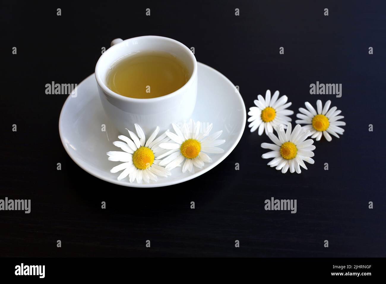 Chamomile tea in a white cup, daisy flowers on saucer. Healing herbal drink on dark wooden table Stock Photo