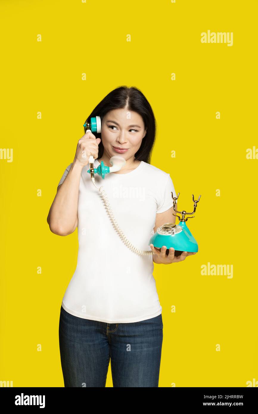 Intrigued middle aged asian woman with vintage, retro telephone in hands interested in, fascinated, draw, lure face expression, isolated on yellow background. Communication concept.  Stock Photo
