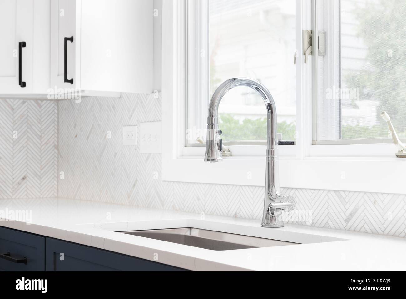 A kitchen sink detail shot with blue and white cabinets, herringbone tile backsplash, and a chrome faucet in front of a window. Stock Photo