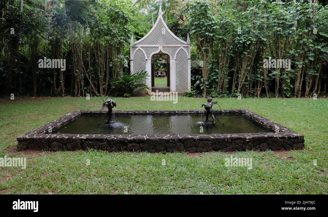 A room in the Allerton Gardens featuring a white gazebo, a wall of trees, a pond with two sculpture fountains surrounded by grass in Kauai, Hawaii Stock Photo