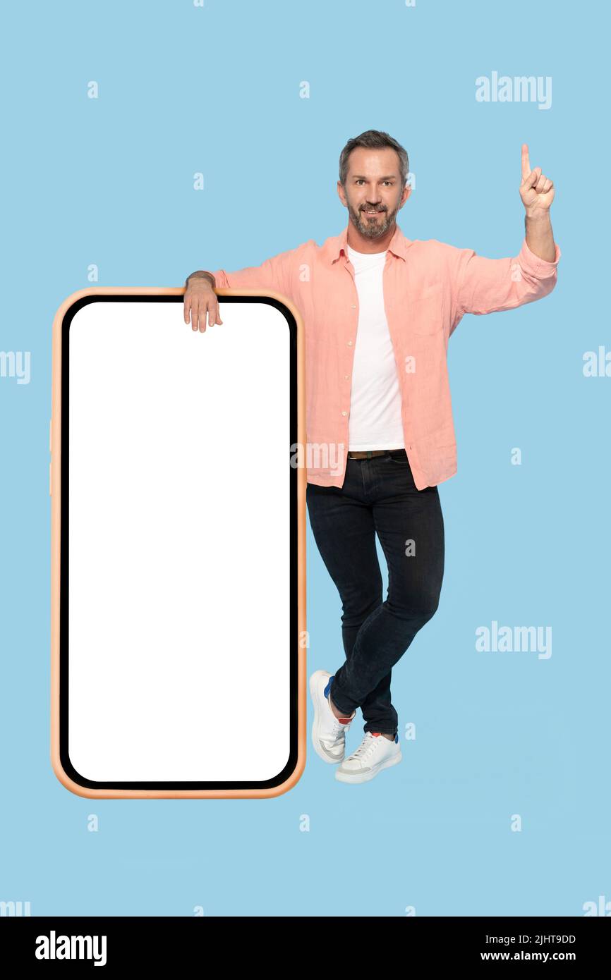 Gesturing attention pointing finger up middle aged grey haired man leaned on huge, giant smartphone with white screen happy smiling on camera wearing peach shirt isolated on blue background. Stock Photo