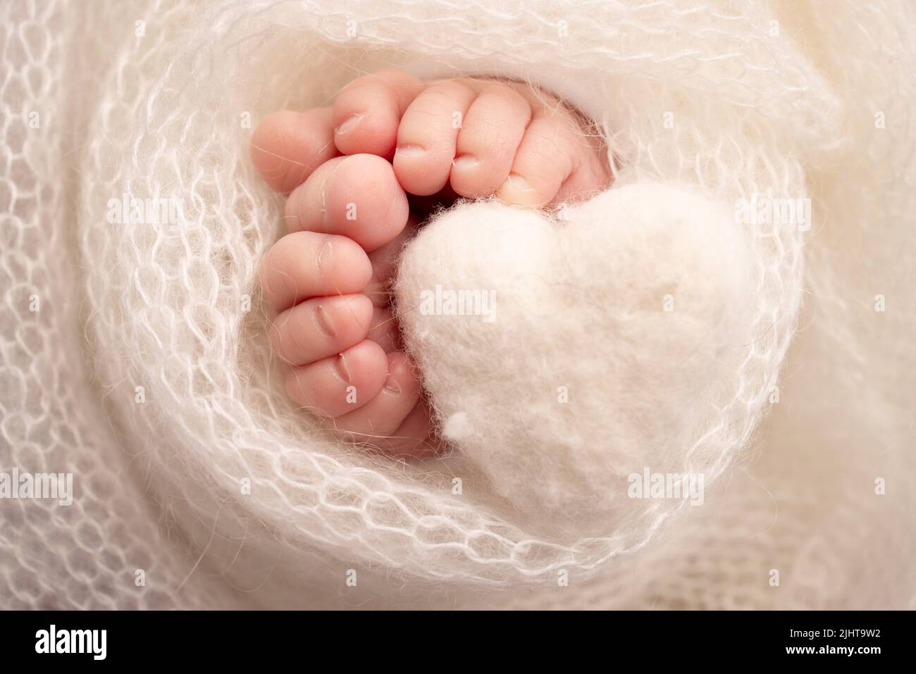 Knitted white heart in the legs of a baby. Close-up of toes, heels and feet of a newborn.  Stock Photo