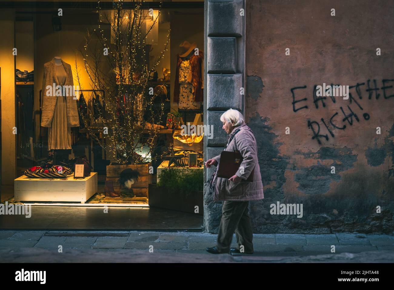 An old lady walking past a fancy fashion shop with graffiti saying 'eat the rich' on the adjoining wall Stock Photo