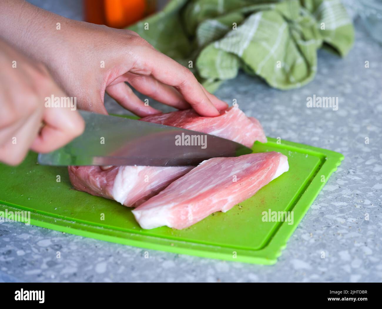 A woman with a knife cutting a raw pork on a green cutting board on a kitchen table. Some pepper and salt are scattered around Stock Photo