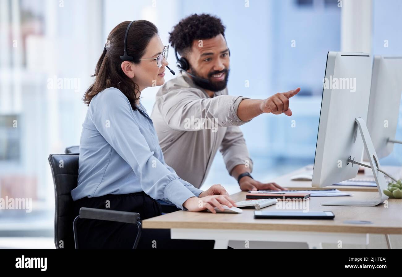 Young caucasian call centre telemarketing agent discussing plans with mixed race colleague on a computer in an office. Two consultants troubleshooting Stock Photo