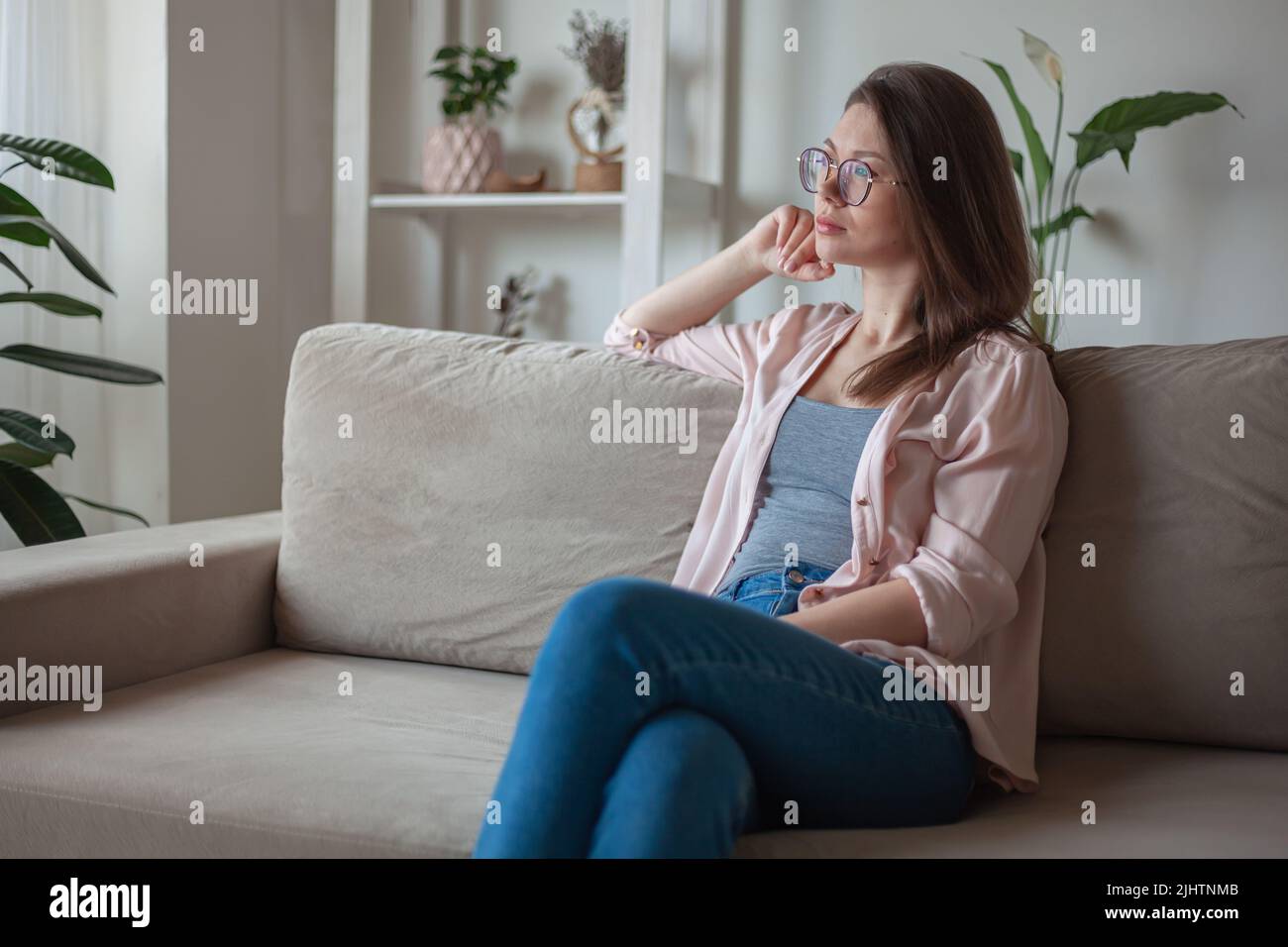 Young woman with eyeglasses sitting on sofa, looking the window Stock Photo