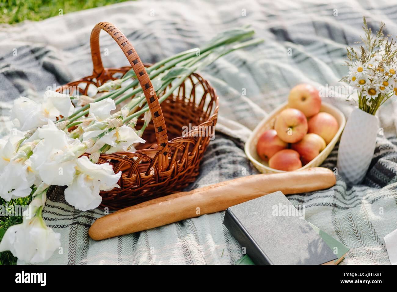 Wicker picnic basket with food and book on gray blanket on grass in ...